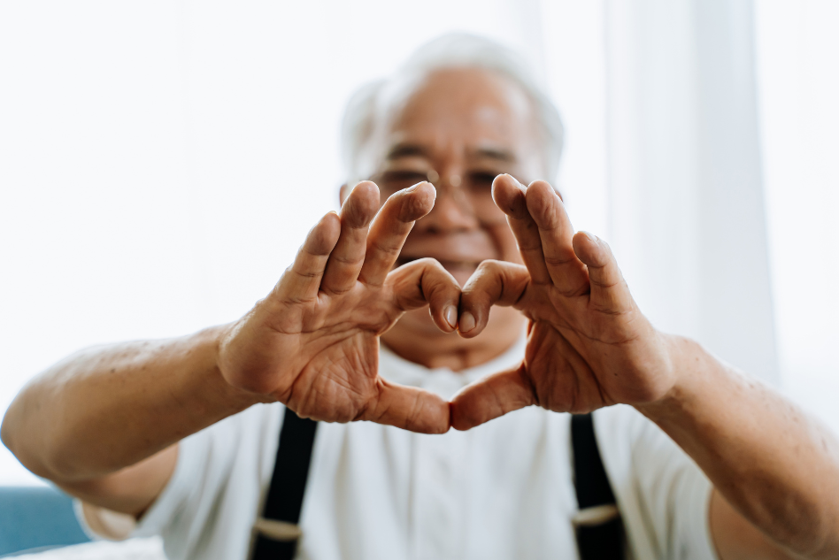 Aged care marketing - An elderly person smiling warmly and forming a heart shape with their hands, symbolizing love and care, against a bright, light-filled background.